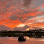 annalee b testimonial image which shows a young woman sitting on a dock with a beautiful orange cloudy sky in the background